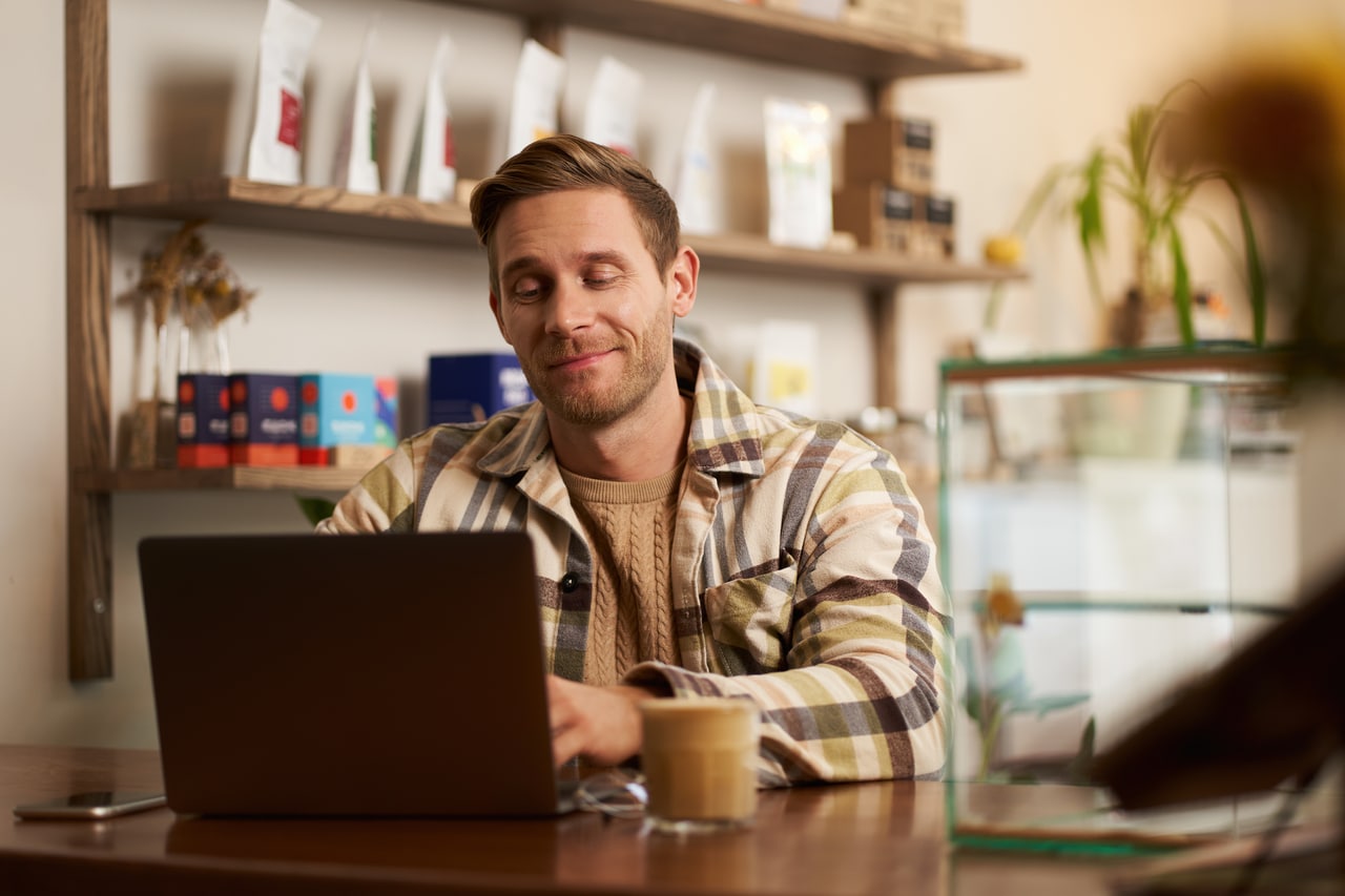 Portrait of handsome young digital nomad, man working in cafe on laptop, looking happy and pleased with his online project, sitting in co-working coffee shop